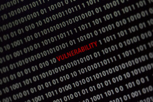 A dark computer screen filled with diagonal rows of white zeros and ones software code, with the word “vulnerability” in bright red and all caps in the middle of the screen.
