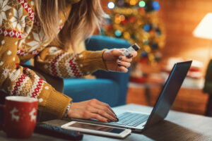Woman in Christmas sweater sitting at her couch online shopping on her laptop. She has a credit card in her hand and a decorated Christmas tree in the background.